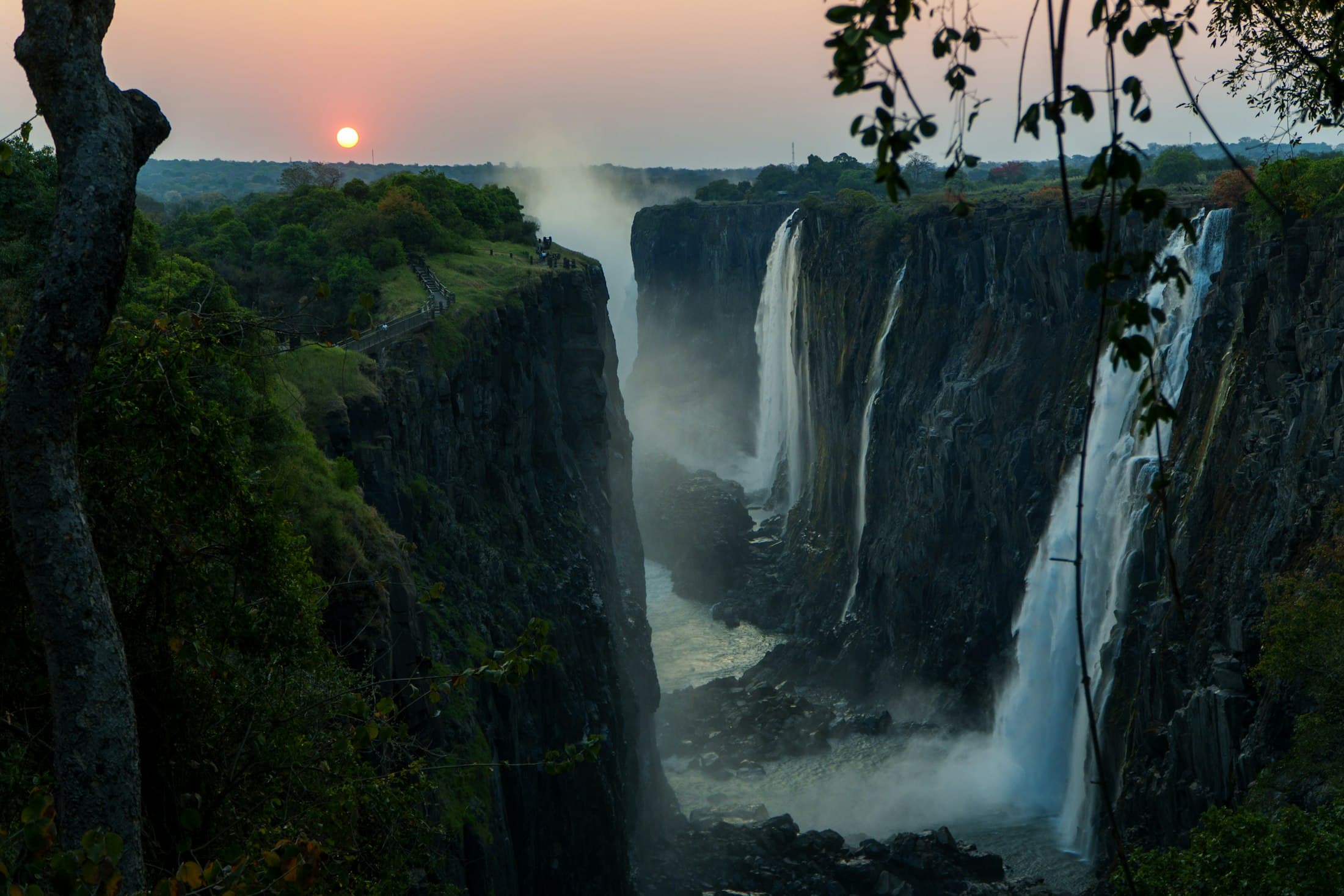 Victoria Falls plunging into a basalt gorge at sunset, with spray rising from the river